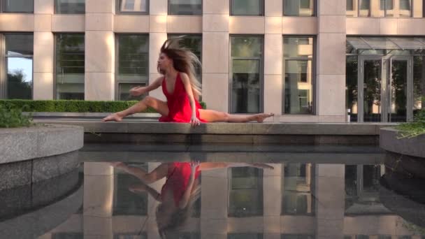Piękna młoda dziewczyna tańczy na ulicy nowoczesnego miasta i odbija się w wodzie. Ma na sobie czerwoną sukienkę. Zwolniony ruch. — Wideo stockowe