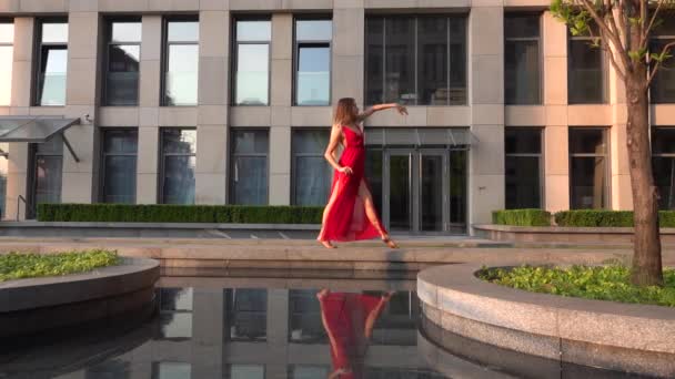 Hermosa joven bailando en la calle de una ciudad moderna y se refleja en el agua. Lleva un vestido rojo. Movimiento lento. — Vídeos de Stock