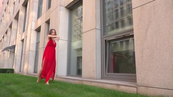 Bella ragazza che balla sull'erba verde sullo sfondo di una città moderna. Indossa un vestito rosso. Rallentatore . — Video Stock