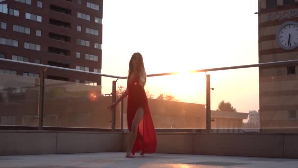 Prachtig jong meisje dansend op de straat van een moderne stad in het zonsondergang licht. Ze draagt een rode jurk. Langzame beweging. — Stockvideo
