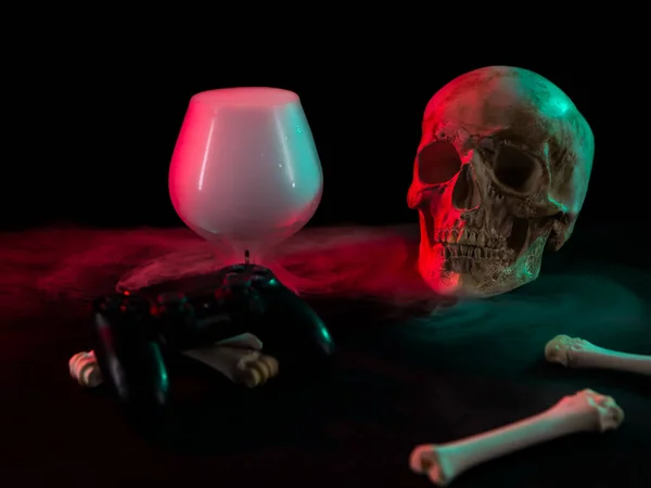 Teschio umano vicino a vetro con fumo denso, ossa e joystick gamepad su sfondo scuro con luce rossa e verde . — Foto Stock