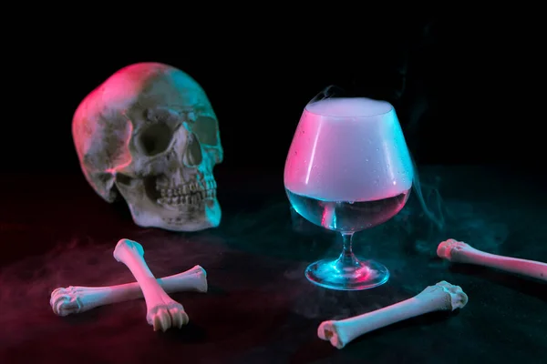 Human skull near glass with thick smoke and bones for Day of the Dead on dark background with red and green light.