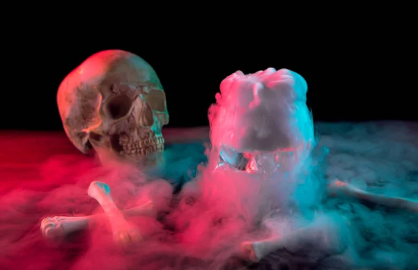 Calavera humana cerca de los huesos y humo blanco grueso que fluye alrededor para el Día de Muertos sobre fondo oscuro con luz roja y verde . — Foto de Stock