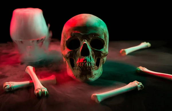 Human skull near glass with thick smoke and bones for Day of the Dead on dark background with red and green light.