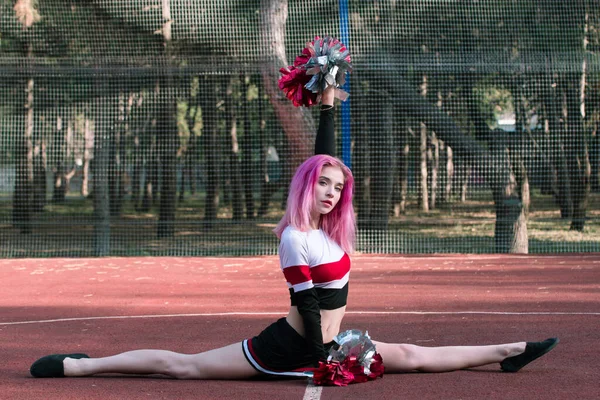 Молода жінка вболіває з рожевим волоссям, сидить у шпагаті і тримає червоні срібні помпони на стадіоні — стокове фото