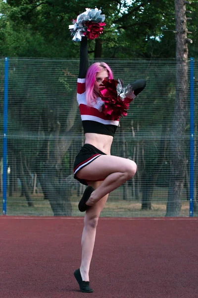Slim fit girl cheerleader with pink hair dancing outdoors with red and silver pompoms at the stadium