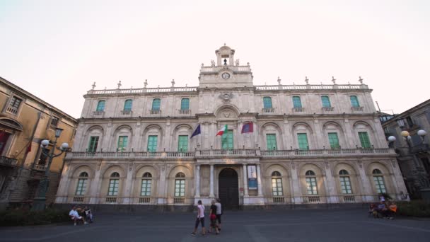 CATANIA, SICILY, ITALIEN - SEPT, 2019: Forntida byggnad av universitetet med flaggor, stor klocka och örn på fasaden. Turister. Blå himmel — Stockvideo