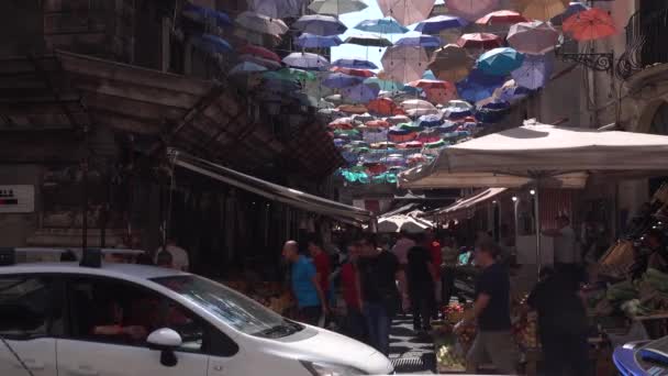 CATANIA, SICILIA, ITALIA - SEPTIEMBRE, 2019: Paraguas coloridos colgando de una cuerda entre edificios antiguos. Bazar callejero, verduras y frutas. Turistas, coches — Vídeos de Stock