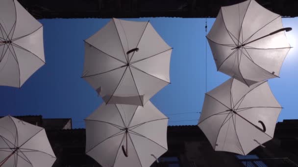 Vita paraplyer hänger på rep mellan gamla byggnader och svajar av vinden. Umbrella Sky Project. Blå himmel, klar sol i Catania, Sicilien, Italien — Stockvideo