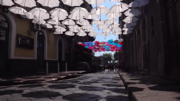 CATANIA, SICILY, ITALIË - SEPT, 2019: Witte en kleurrijke paraplu 's hangend aan touw tussen oude gebouwen. Geparkeerde auto 's. Toeristen lopen door de straat — Stockvideo