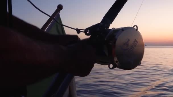 Mãos masculinas fortes que prendem a vara de pesca, carretel de torção. Homem de pesca a partir de iate com bandeira acenando de Itália sobre ele. Mar Mediterrâneo, nascer do sol. Fechar — Vídeo de Stock