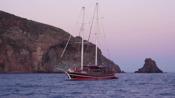 Velero colorido con gente a bordo y barco inflable detrás de él anclado en el mar Mediterráneo contra la isla de Lipari. Sicilia, Italia — Vídeos de Stock