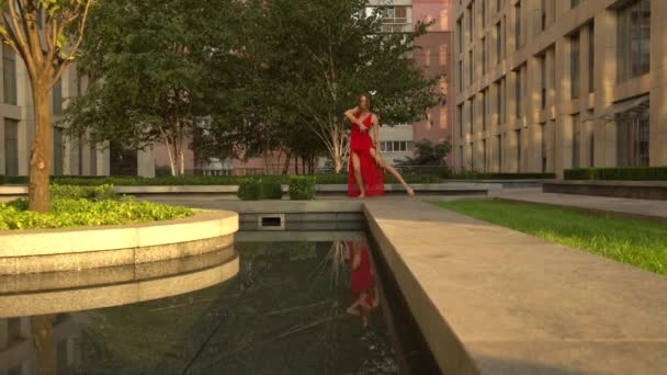 Hermosa joven bailando en la calle de una ciudad moderna y se refleja en el agua. Lleva un vestido rojo. Movimiento lento — Vídeo de stock