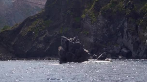 Vågor bryter på liten klippa i havet, berg med byggnad — Stockvideo