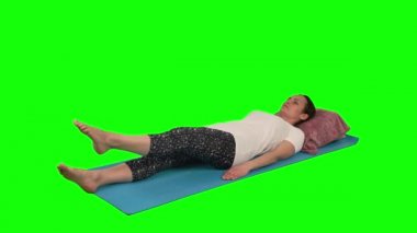 Kadın hasırda yatarken rehabilitasyon egzersizi yapıyor. Stüdyoda yeşil ekrana karşı bacak ve diz kaldırıyor.