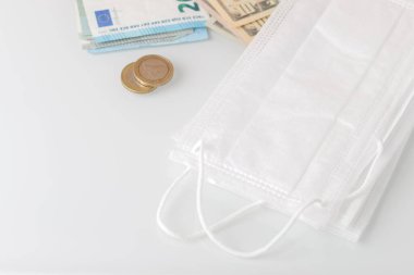 Beyaz bir zemin üzerinde duran bir grup koruyucu tek kullanımlık yüz maskesi dolar, avro, bozuk para ve masraflar konseptiyle birlikte.
