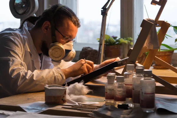 Beyaz önlüklü ve solunum cihazlı usta restoratör ikonu yeniliyor, restoratör restorasyon atölyesinde çalışıyor.