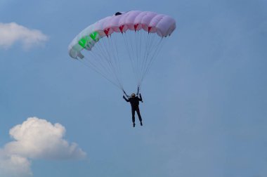 Siyah elbiseli ve siyah gözlüklü bir paraşütçü gökyüzünde beyaz-kırmızı-yeşil bir paraşütle uçuyor.