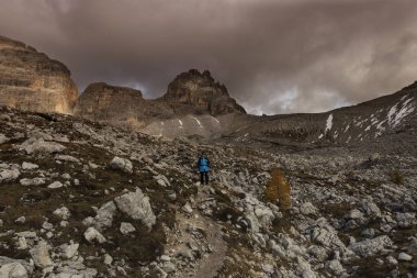 Mavi ceketli bir kadın, bir turist, İtalyan Dolomitleri 'nde Tre Cime Lavaredo' da yürüyüş yapan bir fotoğrafçı, dağları ve arkasında bulutlar olan mavi bir gökyüzü..