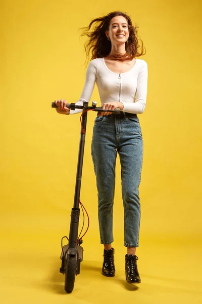 Elektrikli scooterlı güzel bir kız. Sarı arka planda izole edilmiş. Teknoloji ve hareket özgürlüğü.