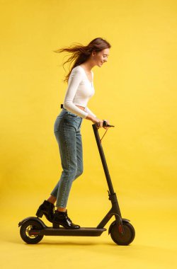 Elektrikli scooterlı güzel bir kız. Sarı arka planda izole edilmiş. Teknoloji ve hareket özgürlüğü.