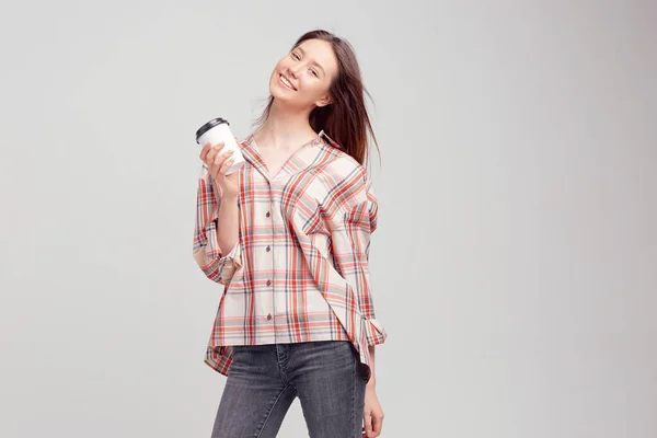 Una jovencita guapa, vestida con ropa casual, sonríe dulcemente y sostiene un vaso de café en un paquete desechable . —  Fotos de Stock