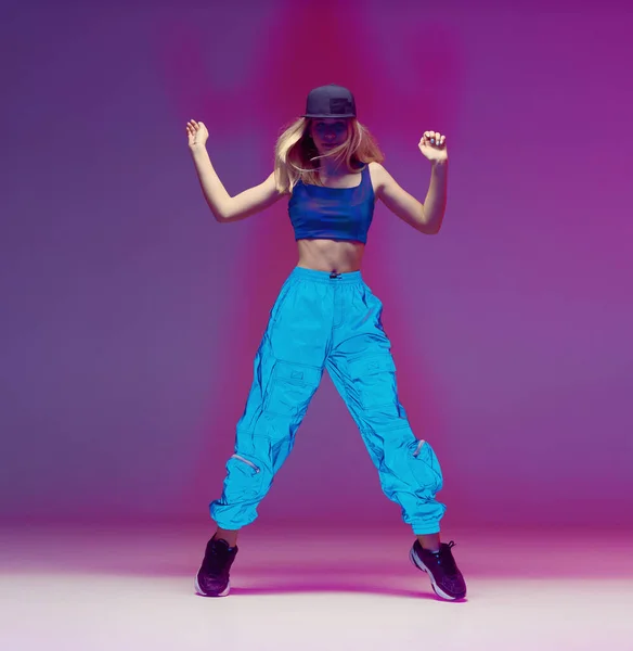 Linda chica adolescente bailando hip hop en pantalones reflectantes, gorra de béisbol, en un estudio con iluminación de neón. Cartel color baile . — Foto de Stock