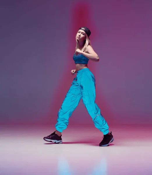 Linda chica adolescente bailando hip hop en pantalones reflectantes, gorra de béisbol, en un estudio con iluminación de neón. Cartel color baile . — Foto de Stock