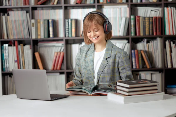 Aranyos fiatal lány diák dolgozik egy egyetemi projekt a könyvtárban egy laptop, egy könyv és fejhallgató. — Stock Fotó