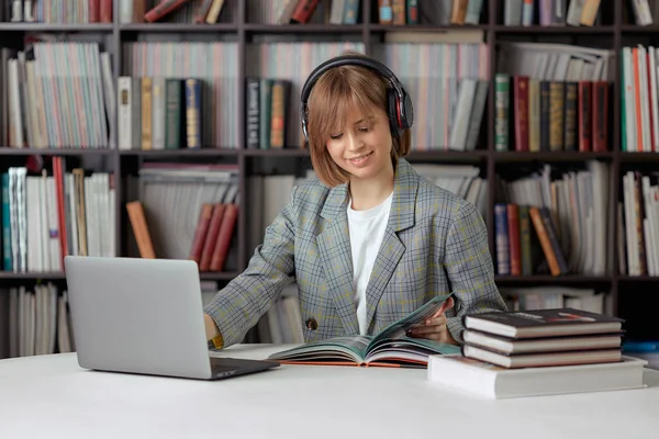 Aranyos fiatal lány diák dolgozik egy egyetemi projekt a könyvtárban egy laptop, egy könyv és fejhallgató. — Stock Fotó