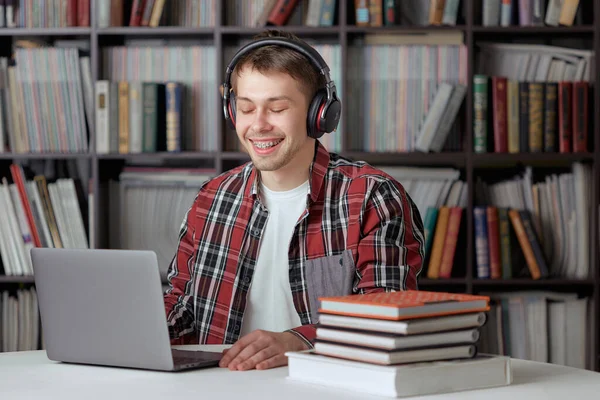 Happy guy listens to a remote audio lesson in the library with wireless headphones and prints a report on a laptop.