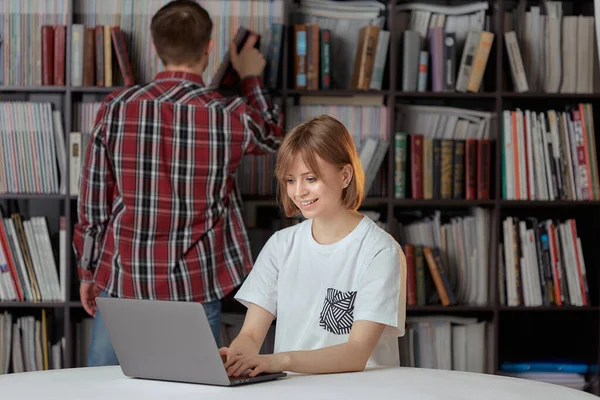 Aranyos fiú és lány diákok dolgoznak egy projekten a városi könyvtárban, könyveket keresnek és jegyzeteket gépelnek egy laptopon. — Stock Fotó