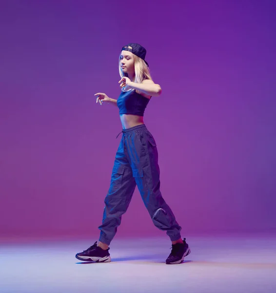Linda chica adolescente bailando hip-hop con ropa elegante, una gorra de béisbol, en un estudio con iluminación de neón. Cartel color baile . — Foto de Stock