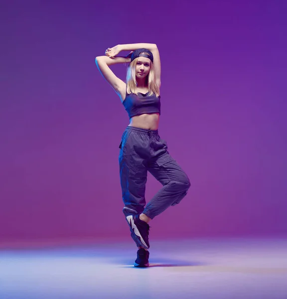 Linda chica adolescente bailando hip-hop con ropa elegante, una gorra de béisbol, en un estudio con iluminación de neón. Cartel color baile . — Foto de Stock