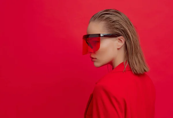 Egy hűvös elegáns lány egy divatos frizura és elegáns szemüveg egy nagy üveg pózol egy élénk piros háttér — Stock Fotó