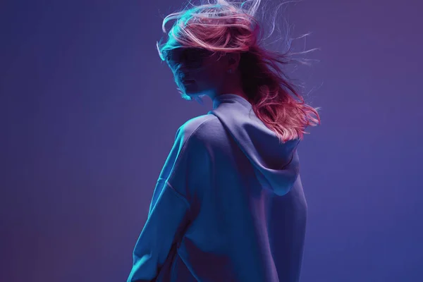 Portré egy elegáns fiatal lány, hűvös pózol kapucnis pulcsi, napszemüveg és a fejlődő haj, a neon háttér. — Stock Fotó