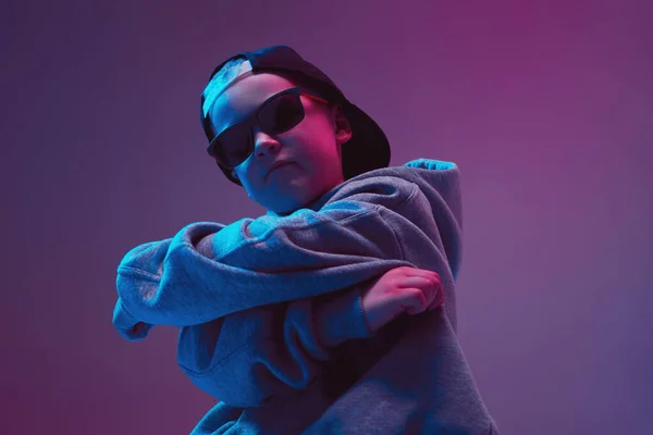 Ritratto di un bambino figo in un'immagine rap, che posa con stile in una felpa con cappuccio, occhiali da sole e un cappuccio su uno sfondo al neon . — Foto Stock