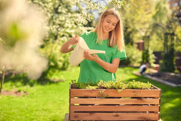 Cute happy teen girl watering flowers, in a wooden pot, from a watering can, in her home garden.