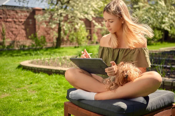 Egy boldog és aranyos tini lány rajzol vázlatokat egy táblára, miközben az udvaron ül egy kutyával, a kertben, a gyepen.. — Stock Fotó
