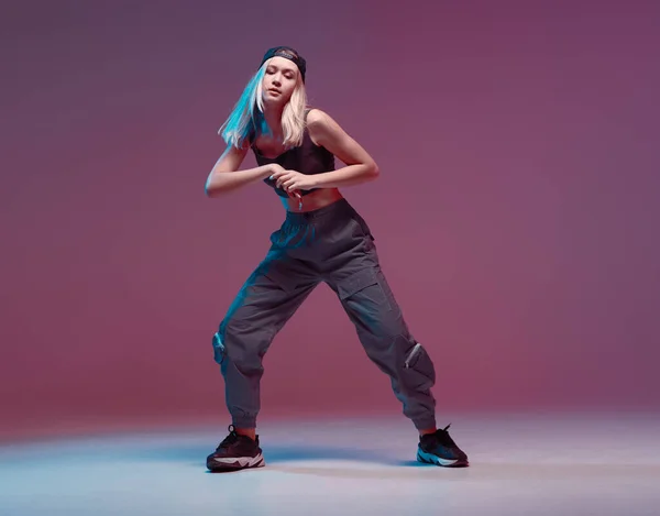 Schattig tienermeisje dansend hiphop in stijlvolle kleding, een honkbalpet, in een studio met neonverlichting. Danskleur poster. — Stockfoto