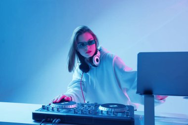 Havalı genç kız DJ karışık konsol ve dizüstü bilgisayarda müziği karıştırıyor, şık kıyafetler içinde, neon arka planda gözlük takıyor..