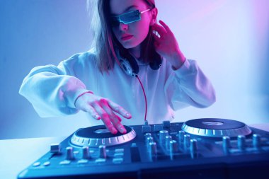 Havalı genç kız DJ karışık konsol ve kulaklıkla müziği karıştırıyor, şık kıyafetler içinde, neon arka planda gözlük takıyor..