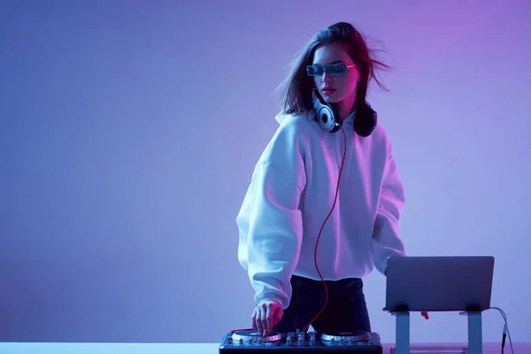 Cool mladá dívka DJ míchá hudbu na mixážní konzoli a notebook, ve stylovém oblečení, brýle na neonovém pozadí. — Stock fotografie