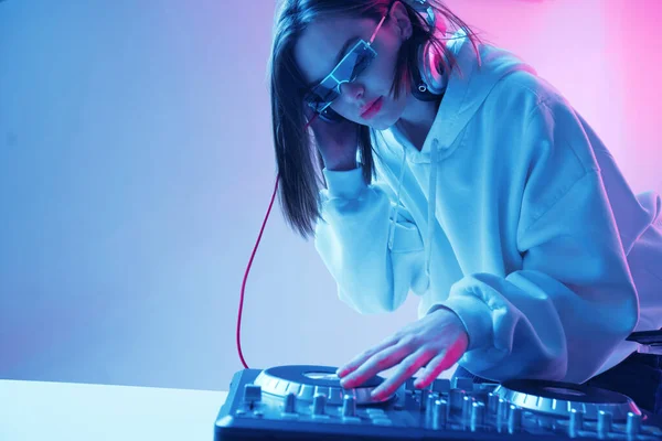 Havalı genç kız DJ karışık konsol ve kulaklıkla müziği karıştırıyor, şık kıyafetler içinde, neon arka planda gözlük takıyor..