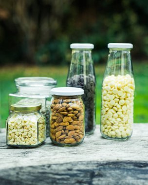 Bademler, fındıklar, tohumlar, yulaflar cam kavanozlarda, doğa arka planında, ahşap bir masada. Sağlıklı gıda kavramları. Vegan kavramı.