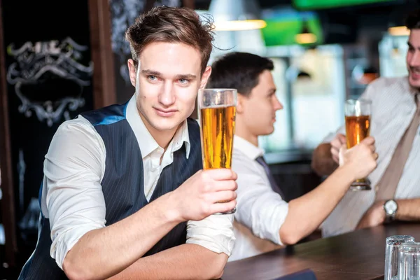Portrait of a young man holding a glass of beer. Three cheerful friends met at the bar and drink a beer while the bartender is standing on the bar. Friends having fun together