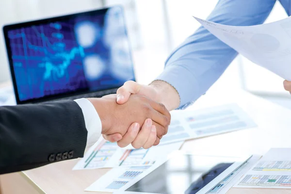 Confident business handshake. Close-up view of a handshake while two successful businessman shaking hands at the table against each in the business office in formal wear and work at a laptop.