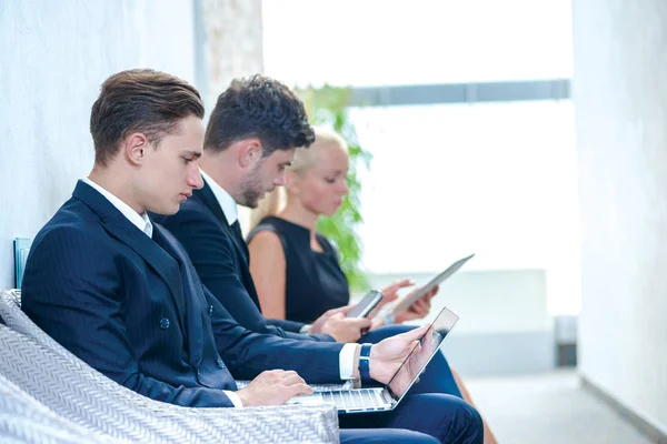 Work at the break. Confident businessman sitting in a waiting room and holding a laptop. Businesspeople sitting in a line and dressed in formal wear.