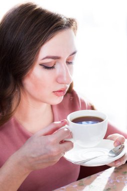 Genç bir kadın pencerenin yakınındaki beyaz bir fincan çay içiyor. 