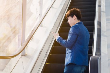 Asyalı yakışıklı iş adamı, yürüyen merdivende dururken gülümsüyor ve akıllı telefon tutuyor. İş ve finans kavramı.